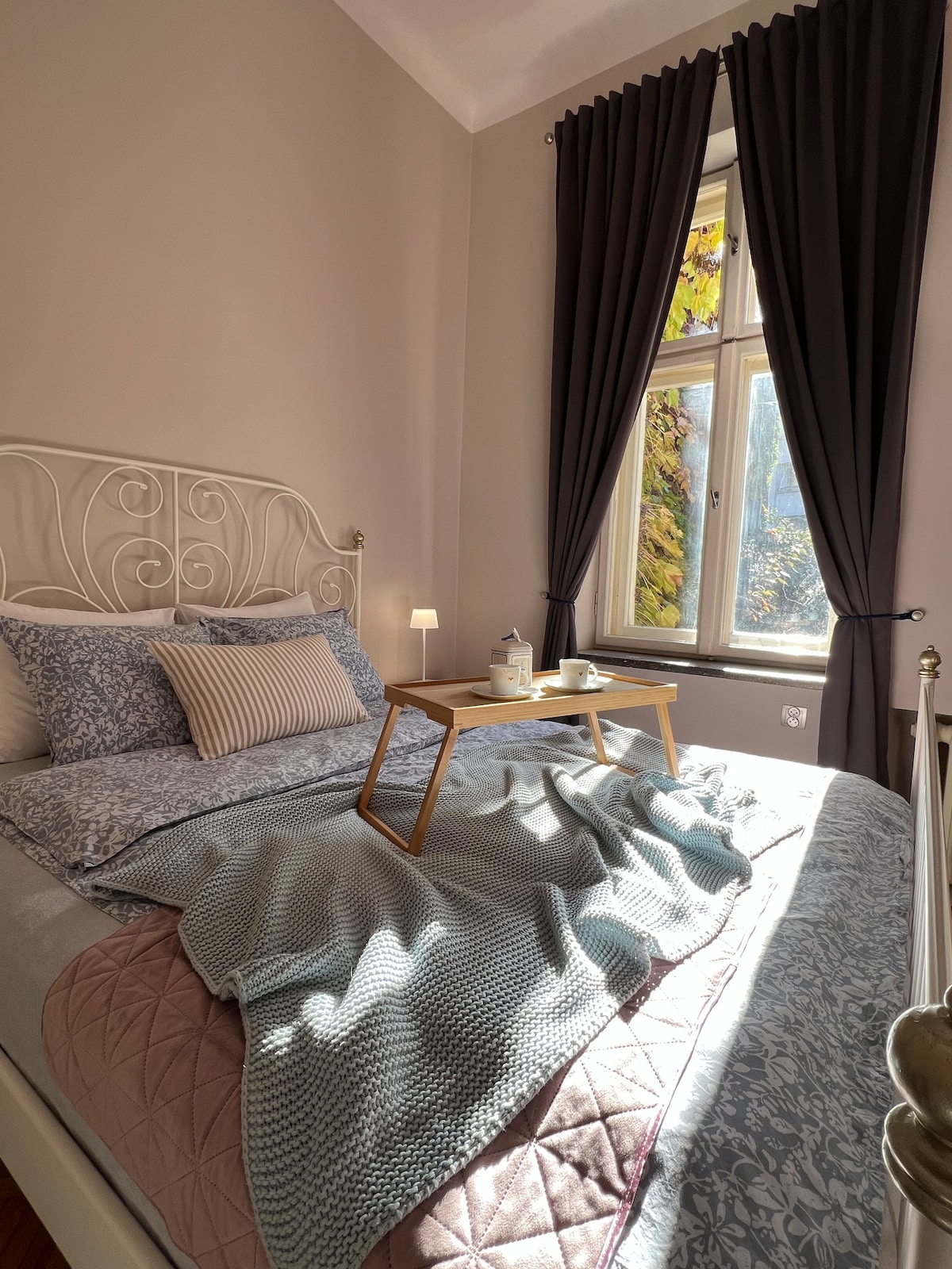 A softly lit bedroom features a spacious bed with layered blankets and decorative pillows. A small tray is set with tea and a teapot, placed on the bed. Large curtains frame a window that lets in natural light, illuminating the tranquil space.