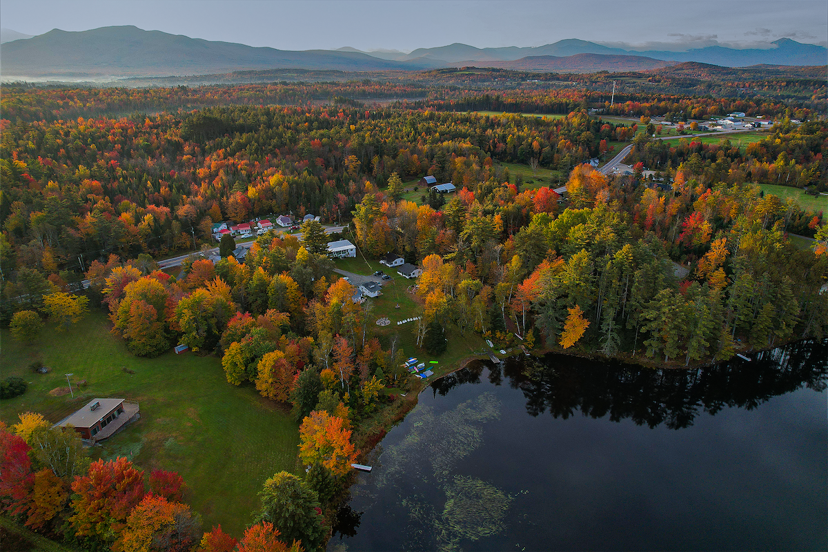 An aerial view captures a vibrant landscape of fall foliage, with rich hues of orange, yellow, and red. A tranquil lake reflects the surrounding trees, while a cluster of buildings and boats dot the shoreline, providing access to outdoor activities amidst the scenic beauty.