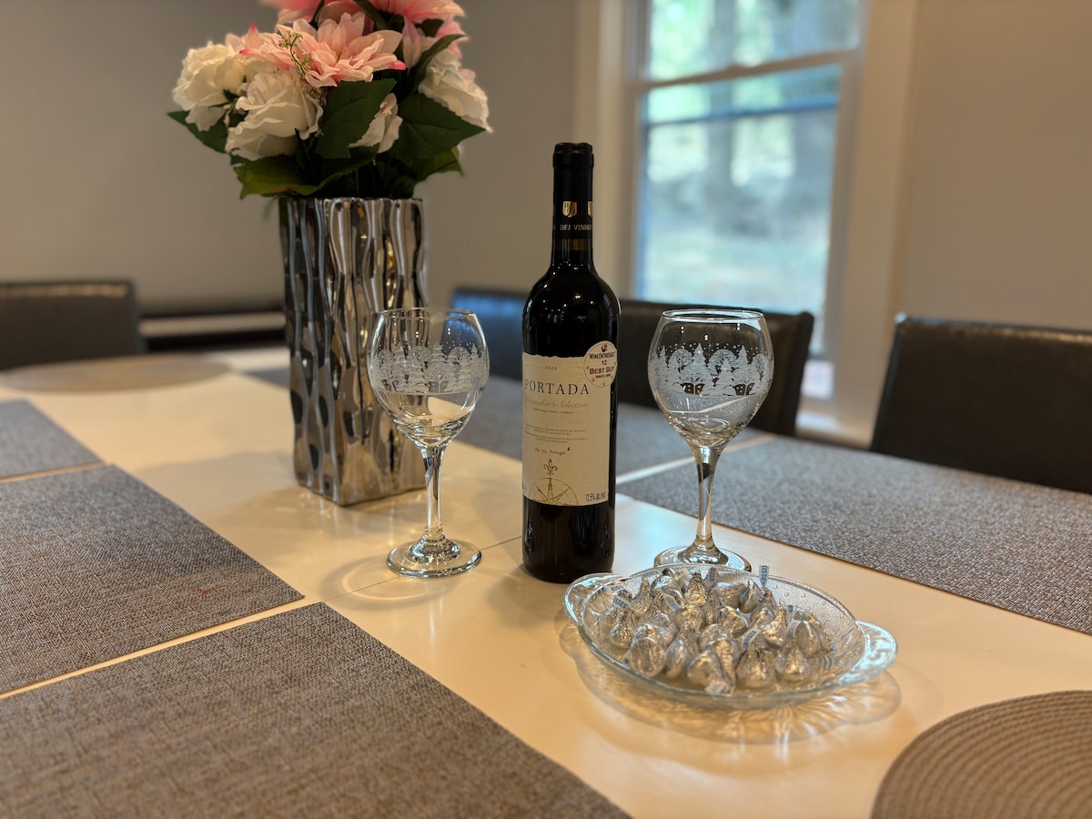 An elegant dining table is set with two crystal wine glasses, a bottle of red wine, and a plate of chocolates. A floral arrangement in a metallic vase adds a touch of freshness. Soft natural light filters in through nearby windows.