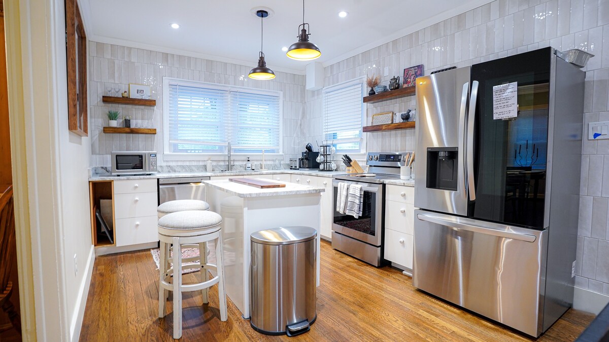 A modern kitchen features stainless steel appliances, including a smart fridge and stove. White cabinetry complements a spacious island with a wooden countertop. Two bar stools are positioned at the island. Natural light enters through the windows, highlighting the tile backsplash and warm wooden flooring.