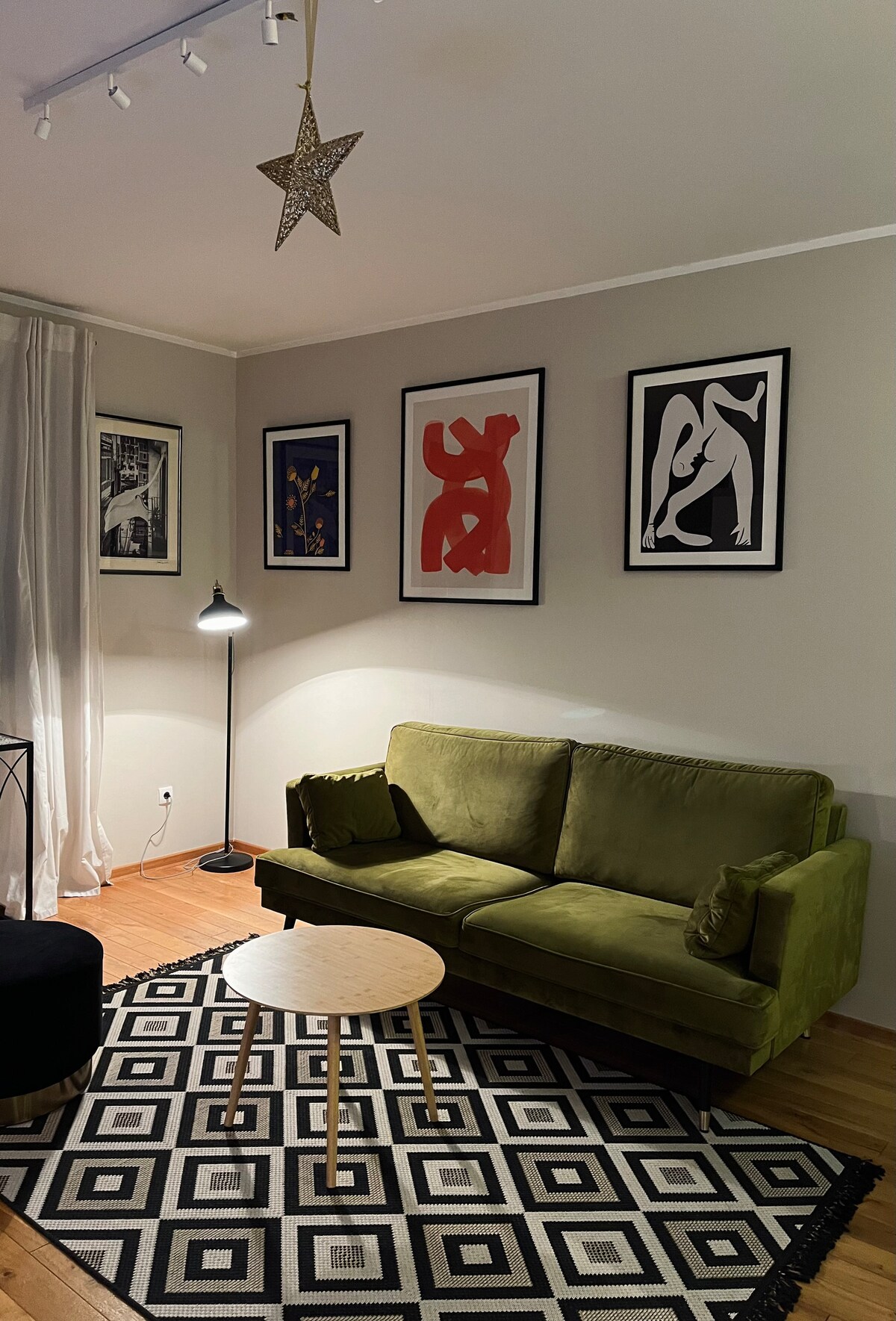 A green sofa is positioned beside a small round table on a black and white patterned rug. The wall is adorned with several framed art pieces, and a standing lamp provides soft lighting, enhancing the warmth of the living space.