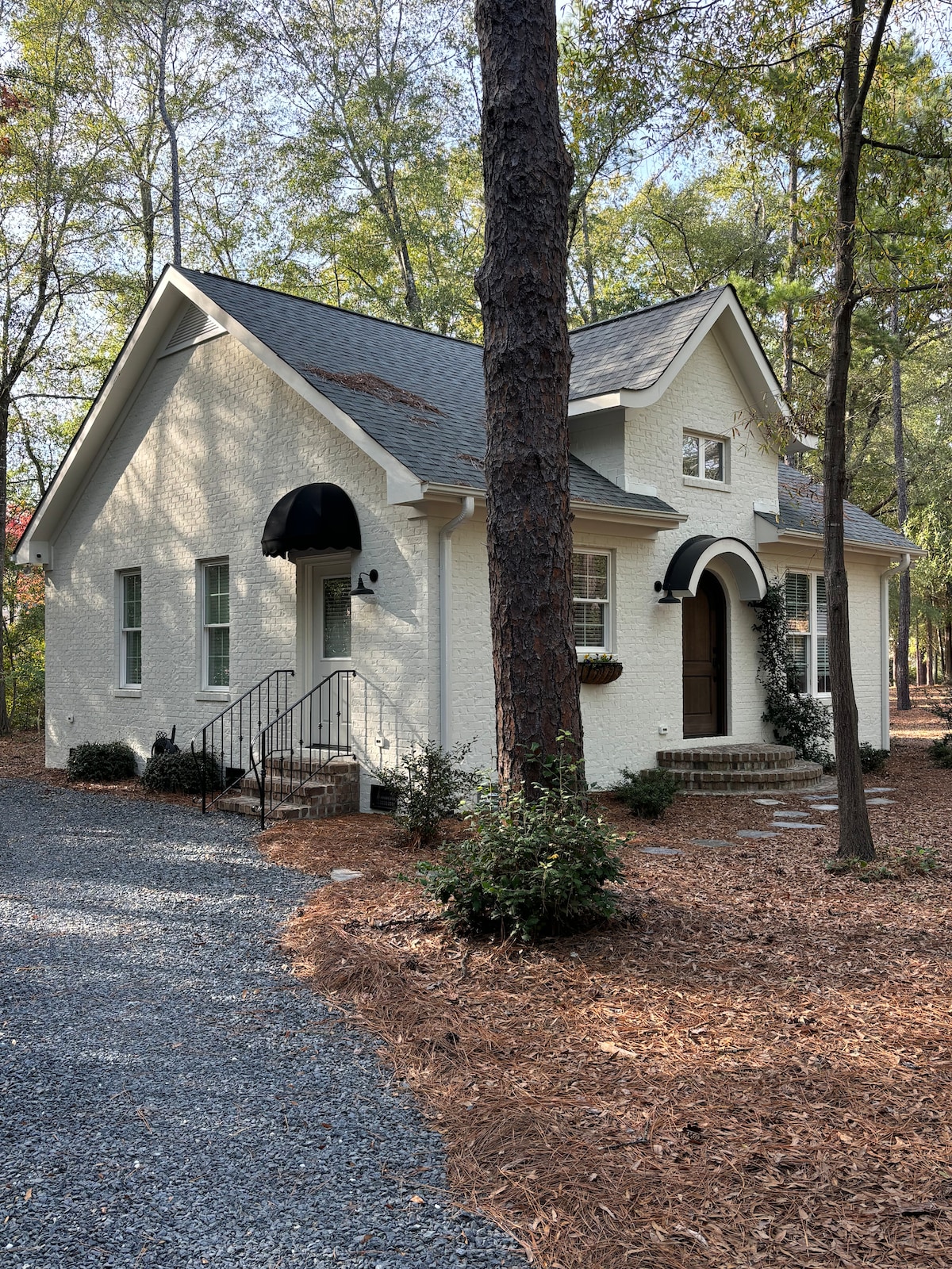 A charming cottage with a white exterior is framed by tall trees and a gravel driveway. Dark shutters and an arched entryway emphasize the architecture. A welcoming staircase leads to the entrance, surrounded by landscaped mulch and bushes.
