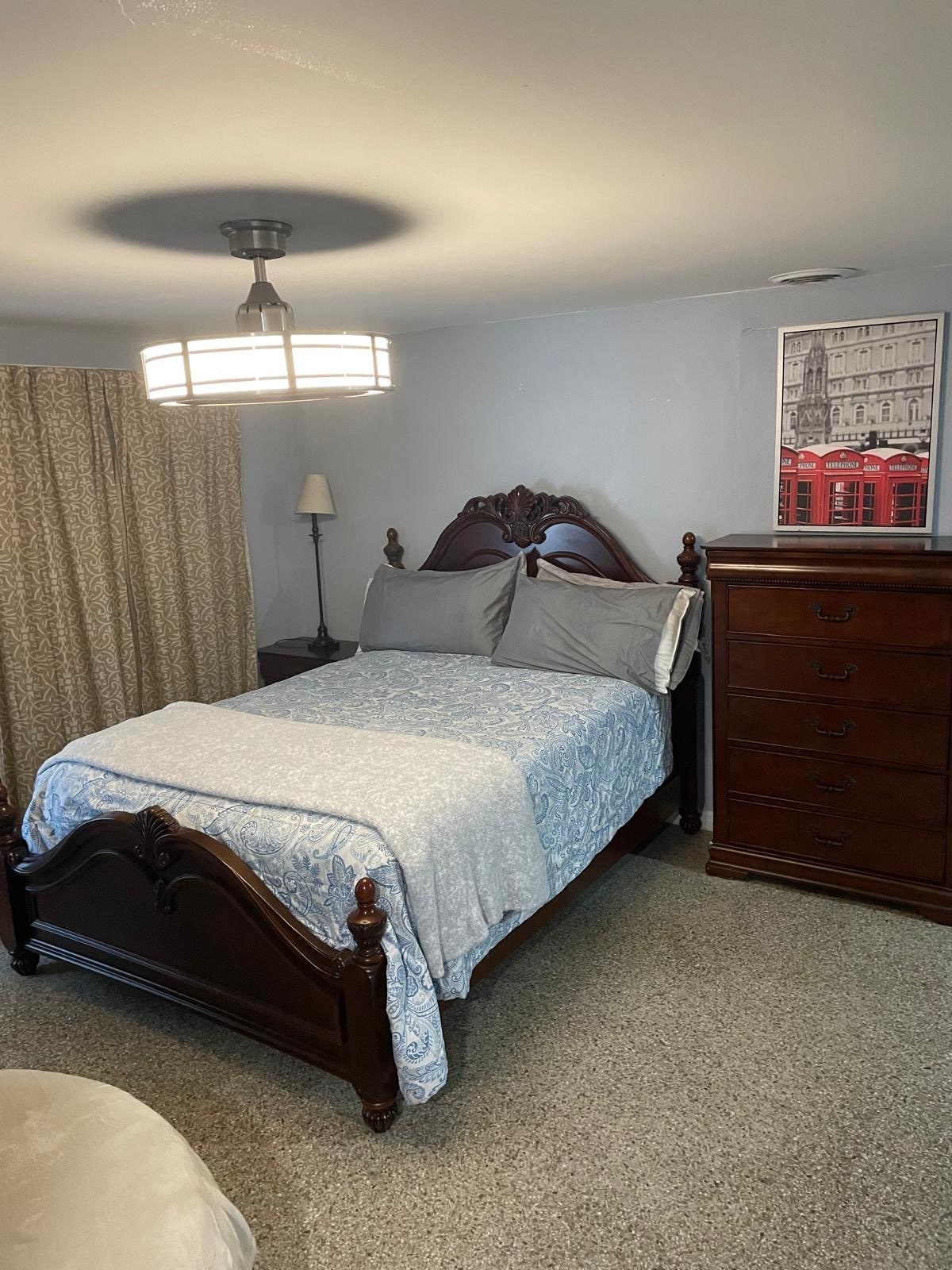 A warm bedroom features a dark wooden bed frame with a light blue patterned blanket over the mattress. A tall dresser stands beside the bed, and a lamp is positioned on a small nightstand. Soft curtains frame a window, while overhead lighting offers a gentle glow.