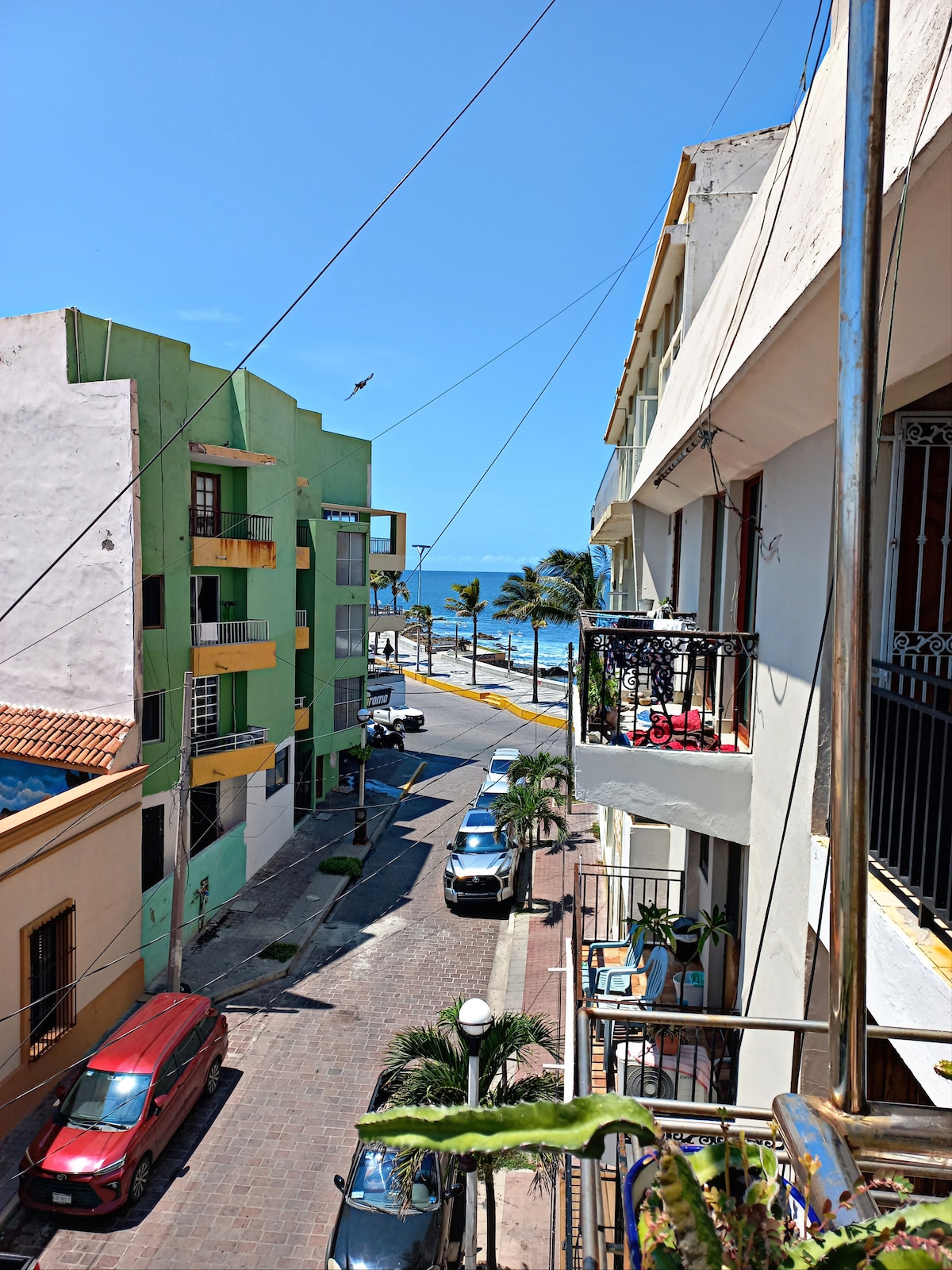A view of a vibrant street lined with colorful buildings, leading to the beach. Palm trees are seen swaying in the breeze, while vehicles are parked along the cobblestone road. The ocean appears in the distance under a clear blue sky.