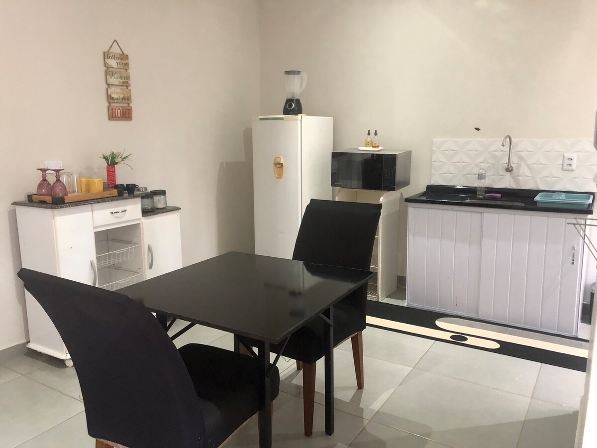 The kitchen area features a black dining table with two chairs, a white refrigerator, and an efficient workspace. Appliances include a blender and a sink with a modern backsplash. The flooring is tiled, and a rug complements the kitchen space.