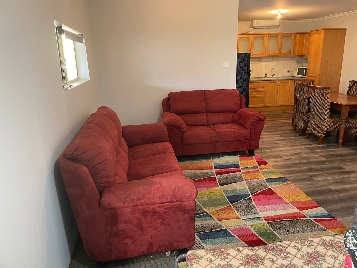 A comfortable lounge area is displayed, featuring two red sofas arranged on a colorful, patterned rug. The space leads to an open kitchen, which is visible in the background, with wooden cabinetry and modern appliances.