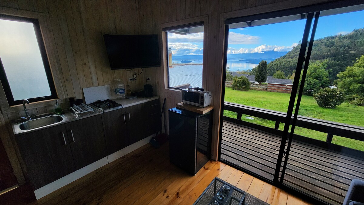 A kitchen area is shown with a sink, stove, and compact refrigerator, accompanied by a wall-mounted TV. Large glass doors provide a view of Lake Ranco and the surrounding landscape, where greenery and distant hills can be seen.