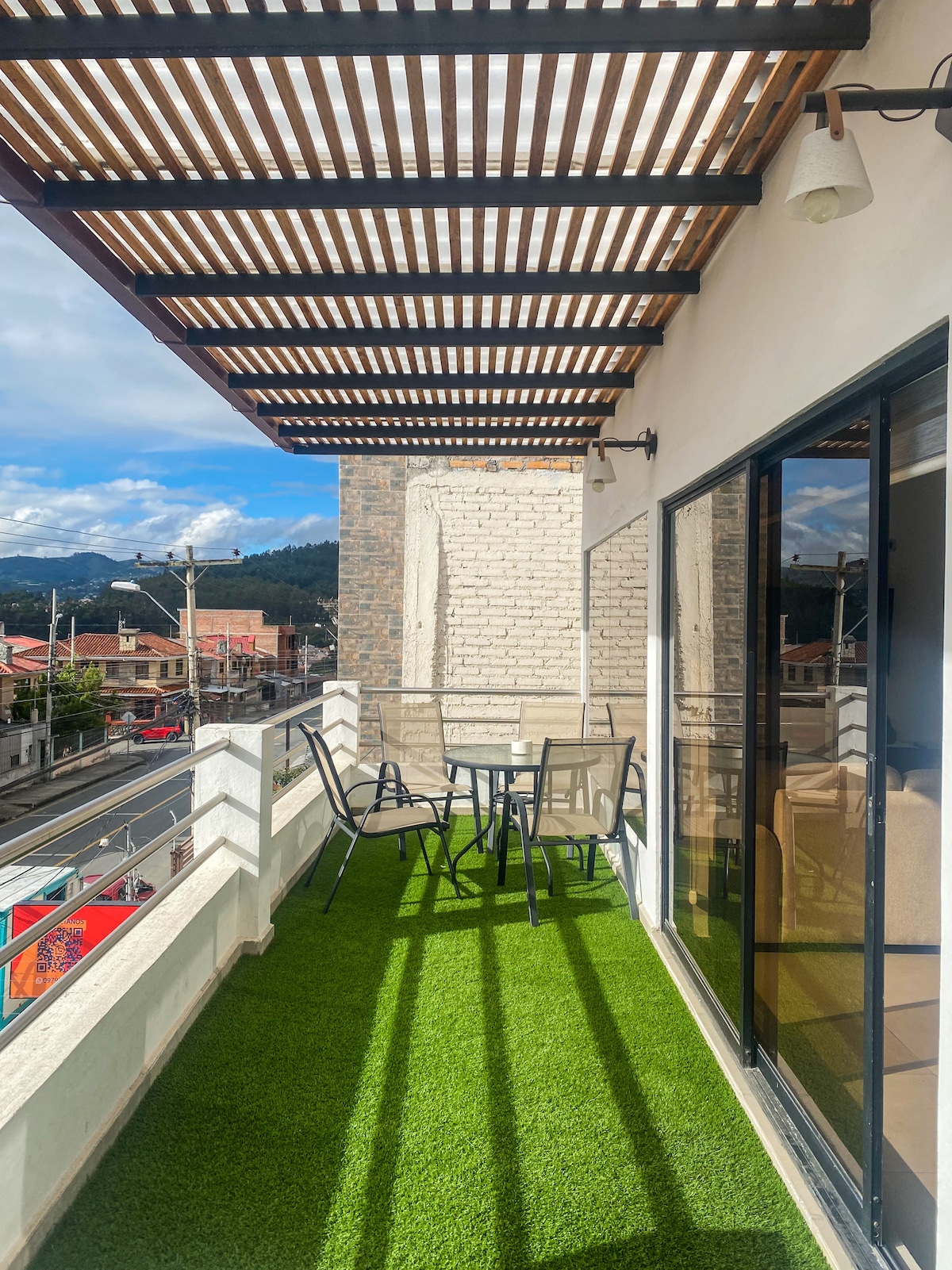 A balcony area is shown featuring artificial grass flooring and a table with two chairs. A pergola provides shade, while large glass doors open towards the interior space. Scenic mountain views and urban surroundings are visible beyond the railing.