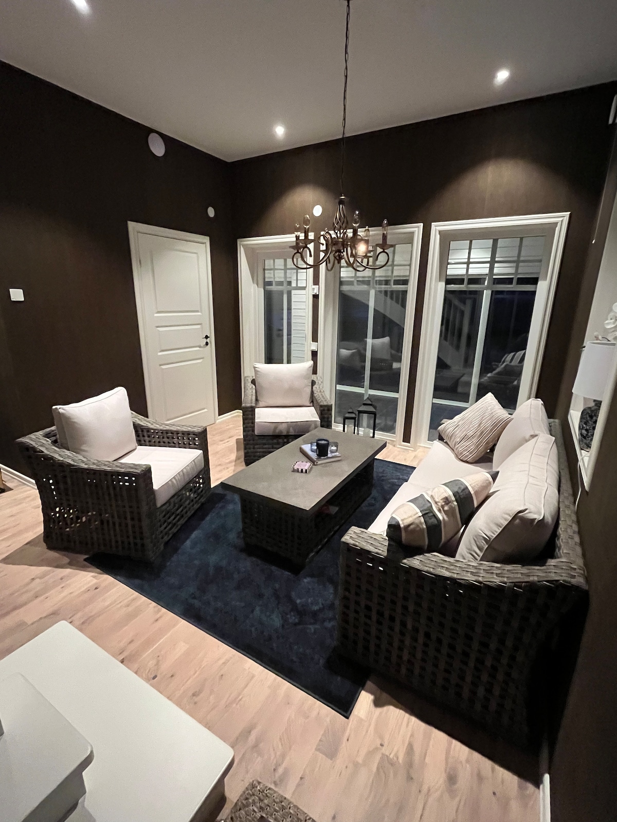 A cozy seating area is arranged with four woven chairs and a central coffee table. Soft cushions add comfort, and large windows offer views of the surrounding area. Subtle lighting creates a welcoming ambiance, highlighting the rich wall color and warm wooden flooring.