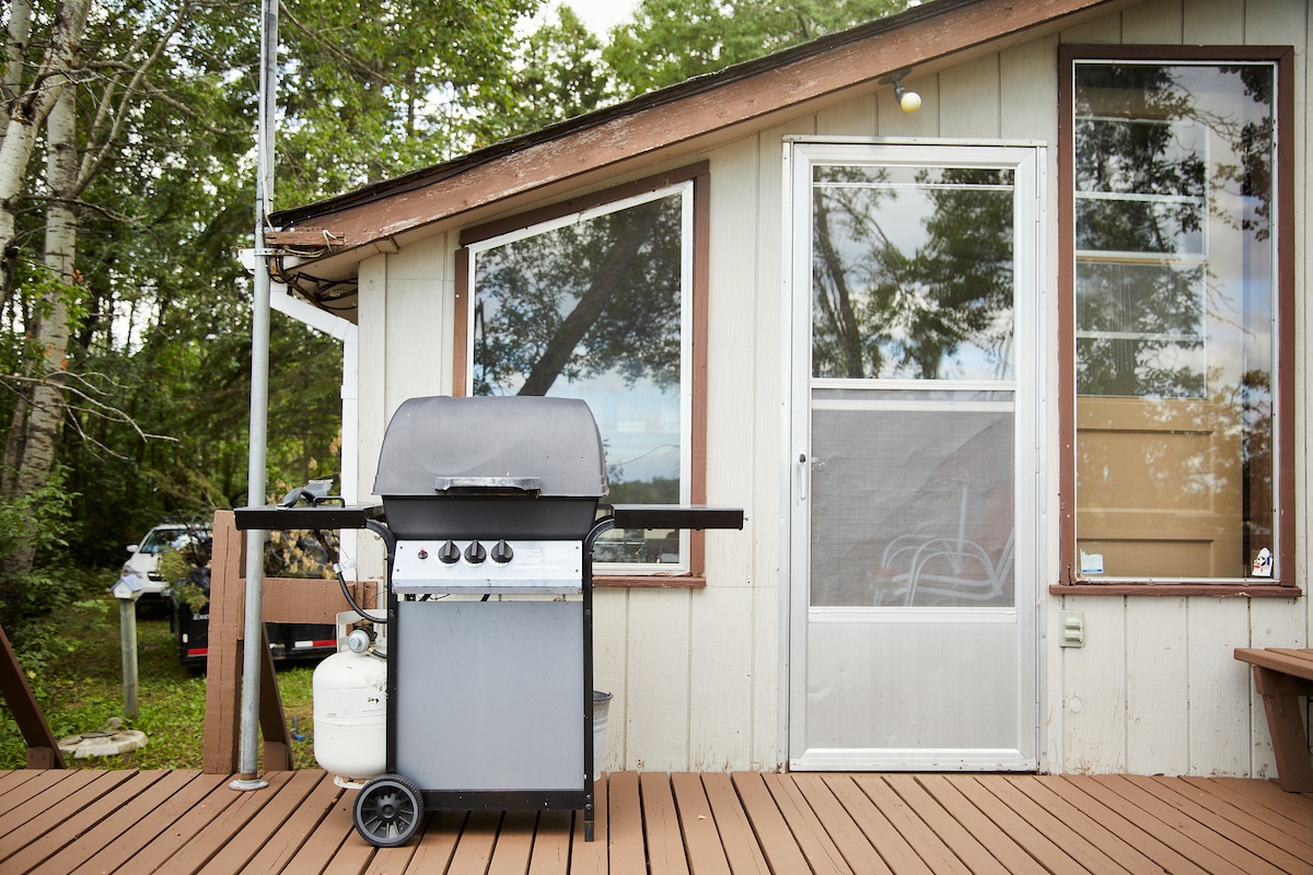 A gas grill sits prominently on the wooden deck, ready for outdoor cooking. The cabin's entrance features large windows that allow natural light to fill the space, blending seamlessly with the surrounding greenery.
