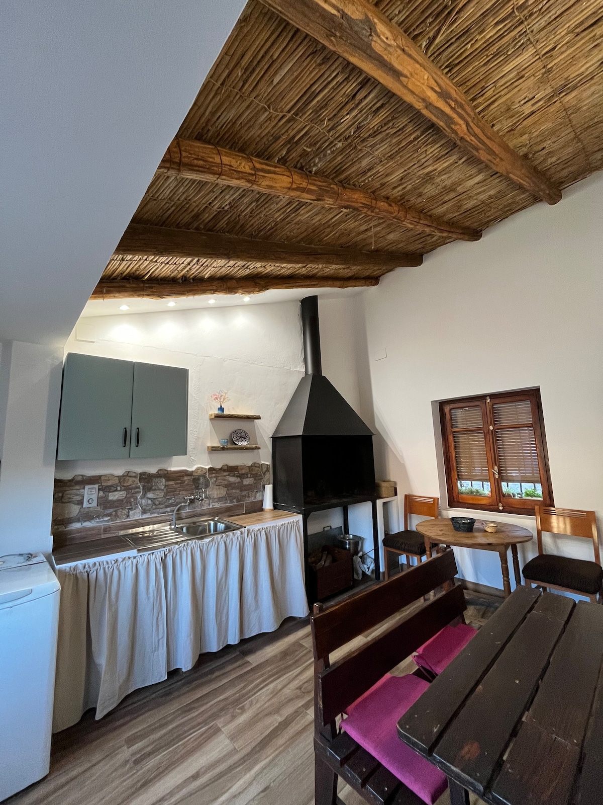 A compact kitchen area features a modern sink and a dark chimney fireplace. Natural wood beams complement the light-colored walls. A small dining table with wooden chairs is positioned nearby, inviting casual meals. Windows adorned with wooden shutters allow soft light to filter in.