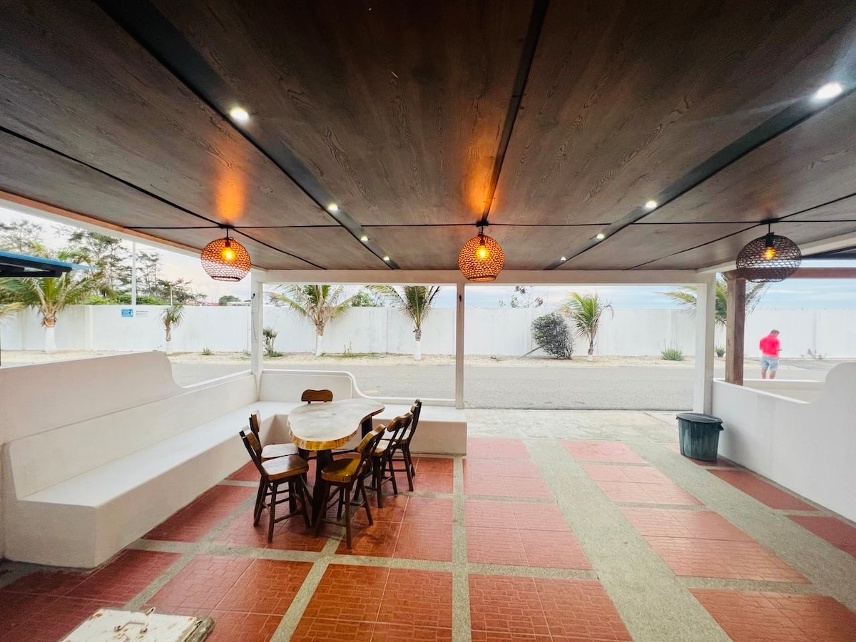 An outdoor seating area features a round table surrounded by wooden chairs. The space is shaded, with a ceiling adorned with pendant lights. Nearby, palm trees are visible, and a glimpse of the beach can be seen in the distance.