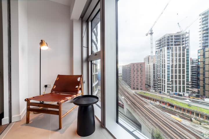 En ljus modern lounge med fönster från golv till tak som visar imponerande utsikt över staden. Utrymmet inkluderar en bekväm soffa och stor TV, vilket gör det perfekt för avkoppling efter en dag i London.