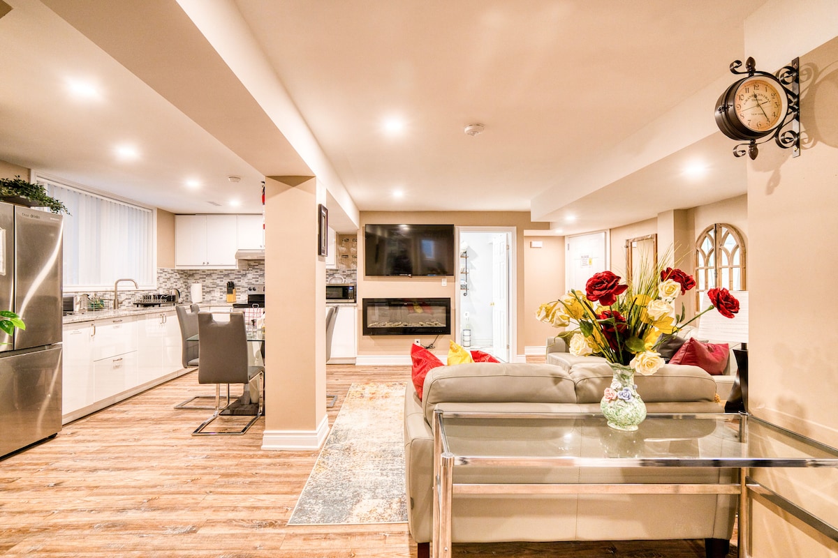 An open-plan living space features modern furnishings, including a light-colored couch adorned with decorative flowers. A sleek kitchen is visible in the background, equipped with stainless steel appliances. Soft lighting highlights the wood-like flooring, contributing to a warm, inviting ambiance.