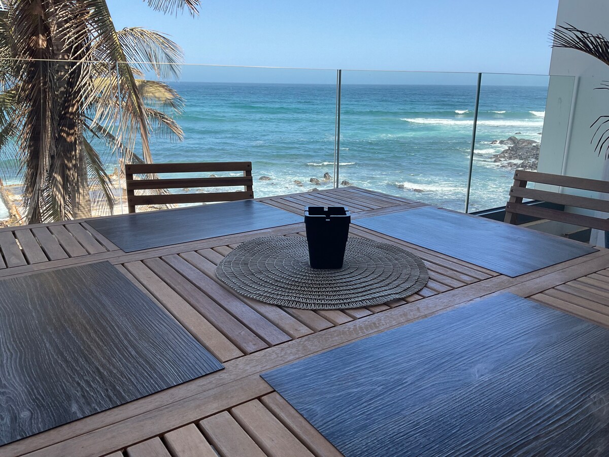 A wooden dining table is set on a terrace, overlooking the ocean. The table is accompanied by several wooden chairs. A round woven placemat sits at the center, featuring a small black container. Palm trees frame the view of the waves and clear blue sky.
