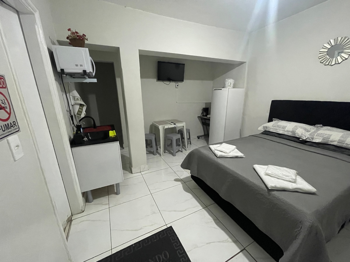 A well-furnished flat features a spacious bed covered with neutral-toned linens. A small dining area with a table and stools is visible. A kitchenette with essential appliances, including a fridge and microwave, is present. A wall-mounted TV adds entertainment options.