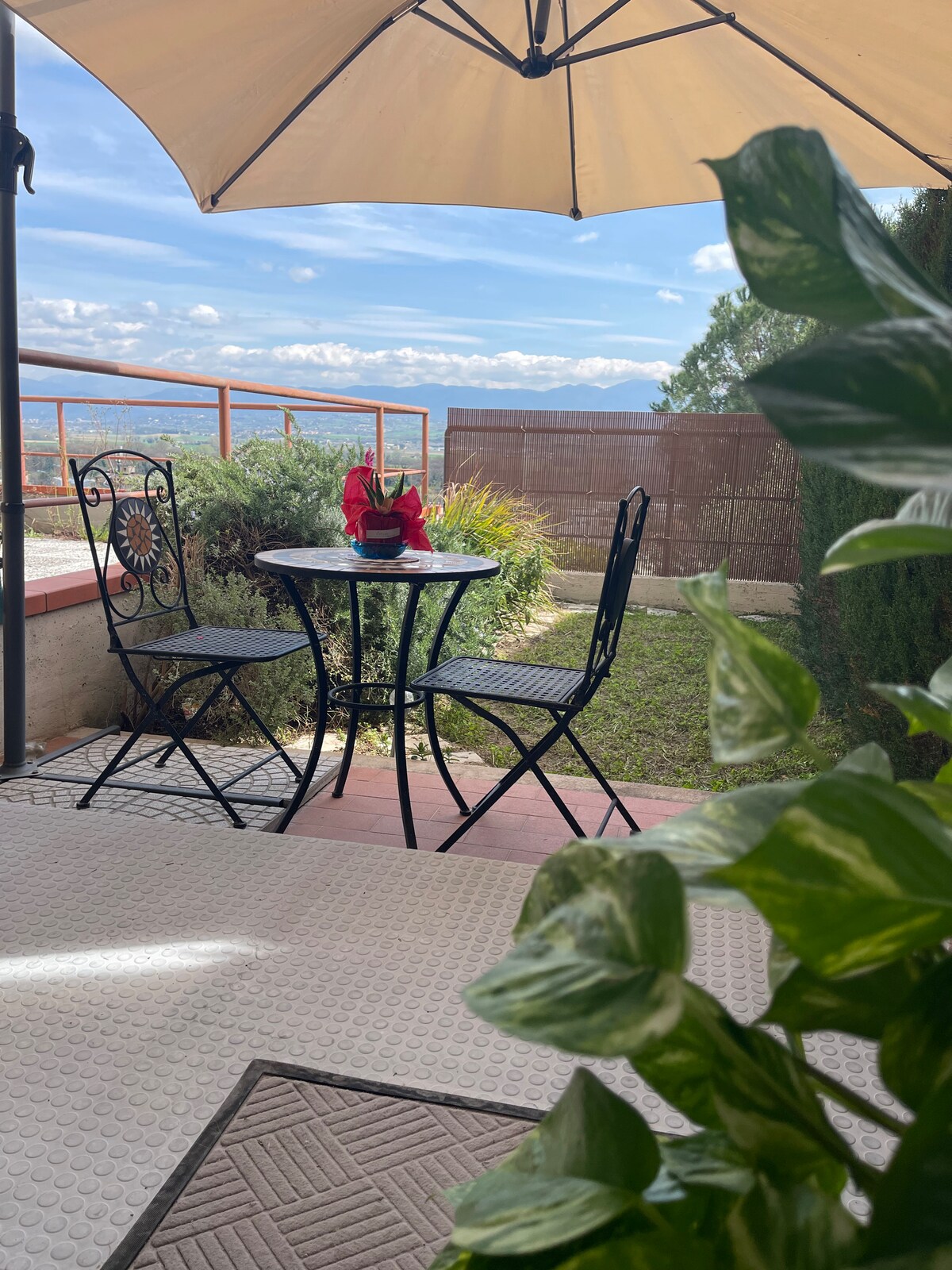 An outdoor seating area is featured, consisting of a small black table with two chairs under a large umbrella. A potted plant decorates the table, and greenery is visible in the background, providing a serene view of the surrounding landscape.