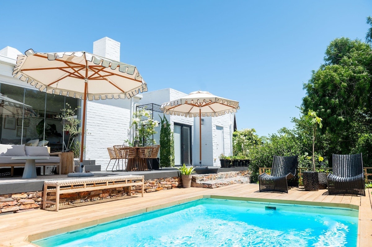 A refreshing pool surrounded by wooden decking is highlighted by two large umbrellas. Comfortable seating areas with black wicker chairs offer shaded spots for relaxation. Lush greenery and well-maintained outdoor spaces enhance the serene setting, inviting moments of leisure and family fun.