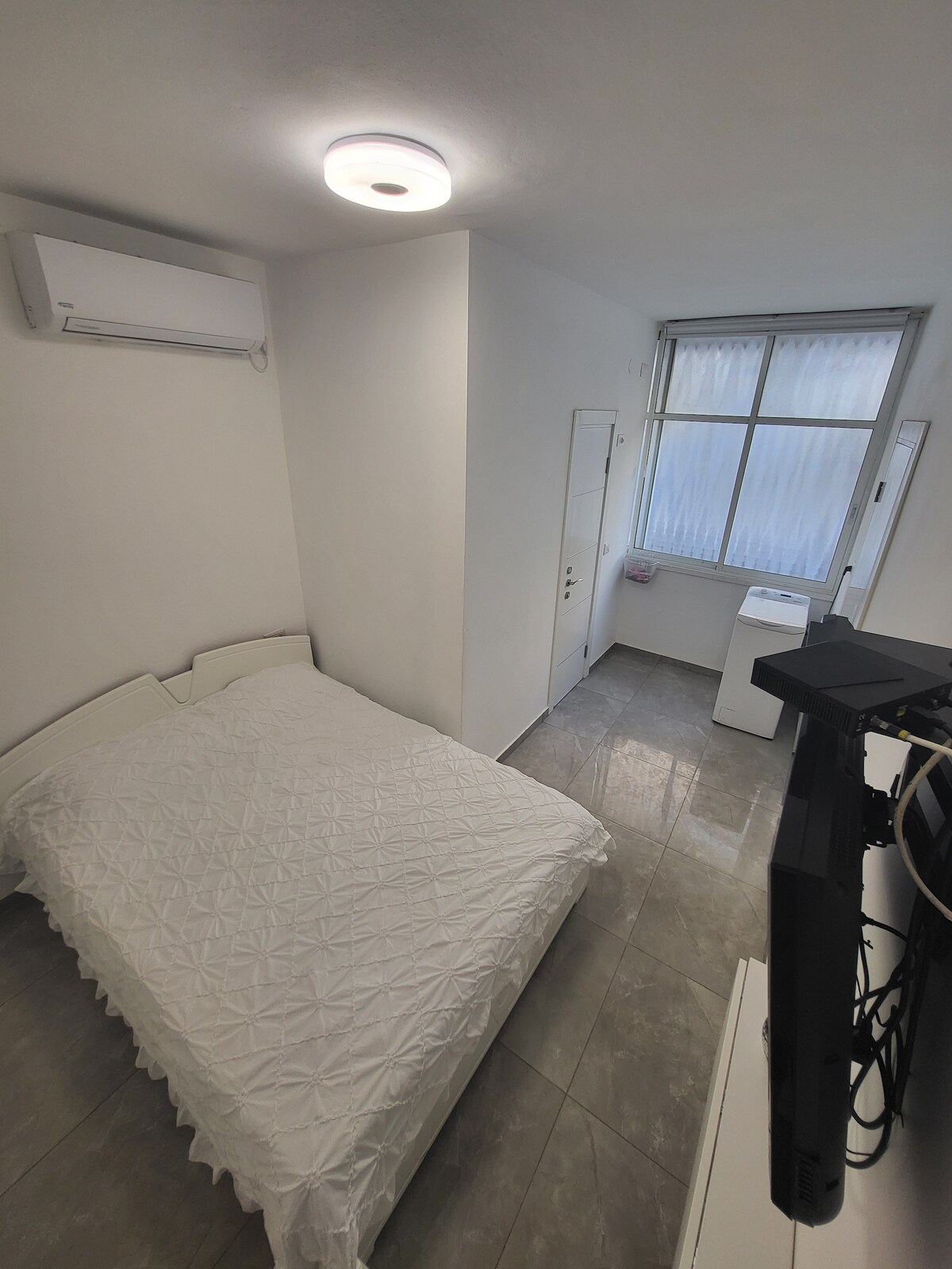 A bright sleeping area features a spacious bed with a textured white coverlet. A window allows natural light to enter, illuminating the room's neutral walls. An air conditioning unit is mounted above, while a washing machine is visible in the corner near the entrance door.