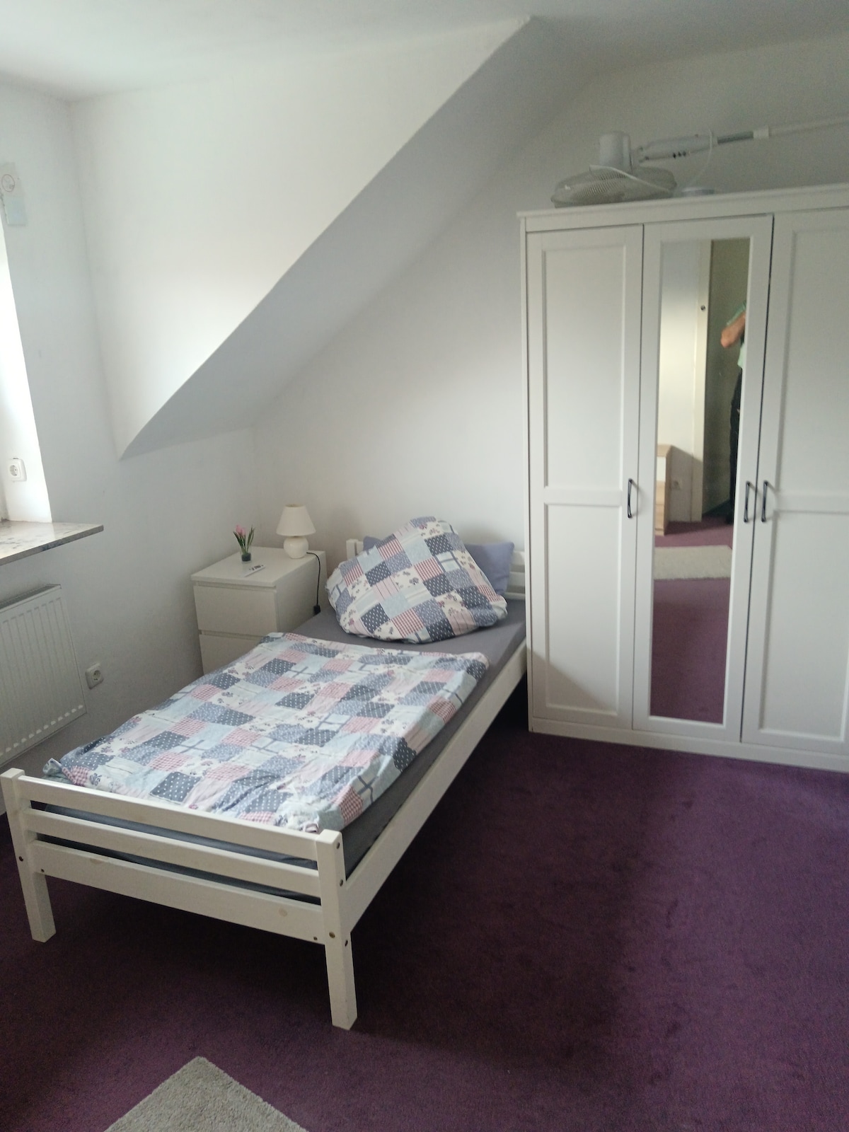A cozy guest room is presented with a single bed featuring a patterned quilt. A small bedside lamp and white storage cabinet are positioned beside the bed. A mirrored wardrobe reflects the room's design, while soft purple carpeting adds a gentle touch to the space.