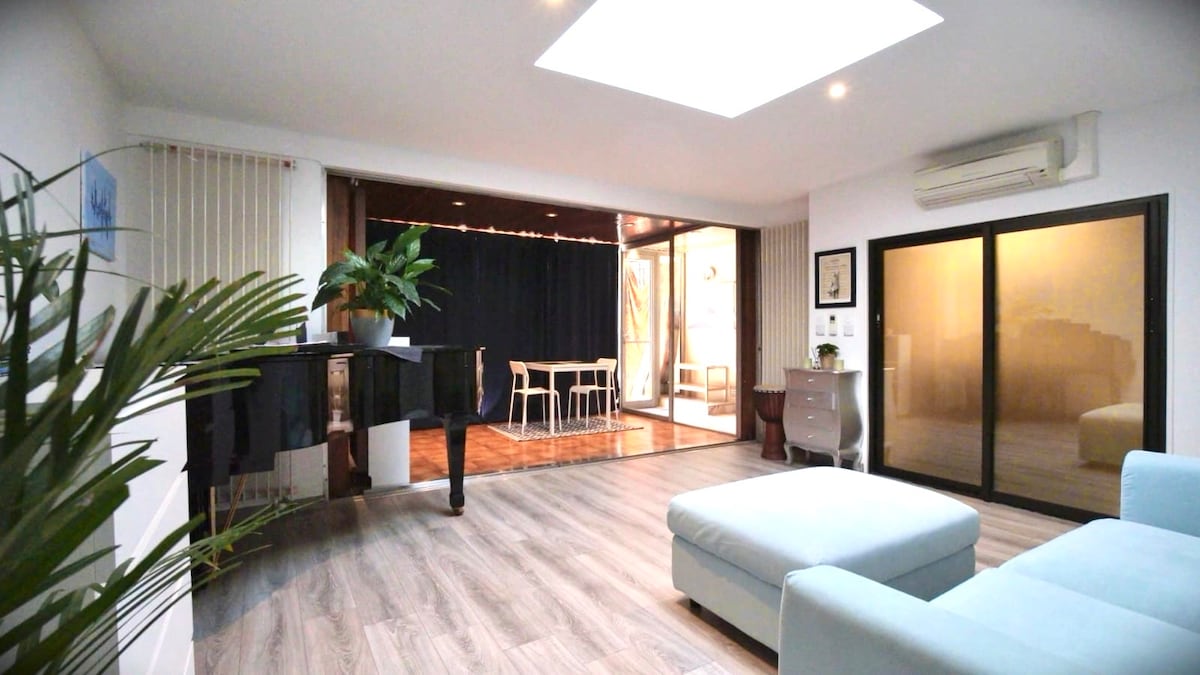A spacious studio is shown with abundant natural light filtering through a skylight and large glass doors. A grand piano occupies one corner, while a comfortable seating area is visible. Light-colored wooden flooring complements the serene ambiance created by greenery.