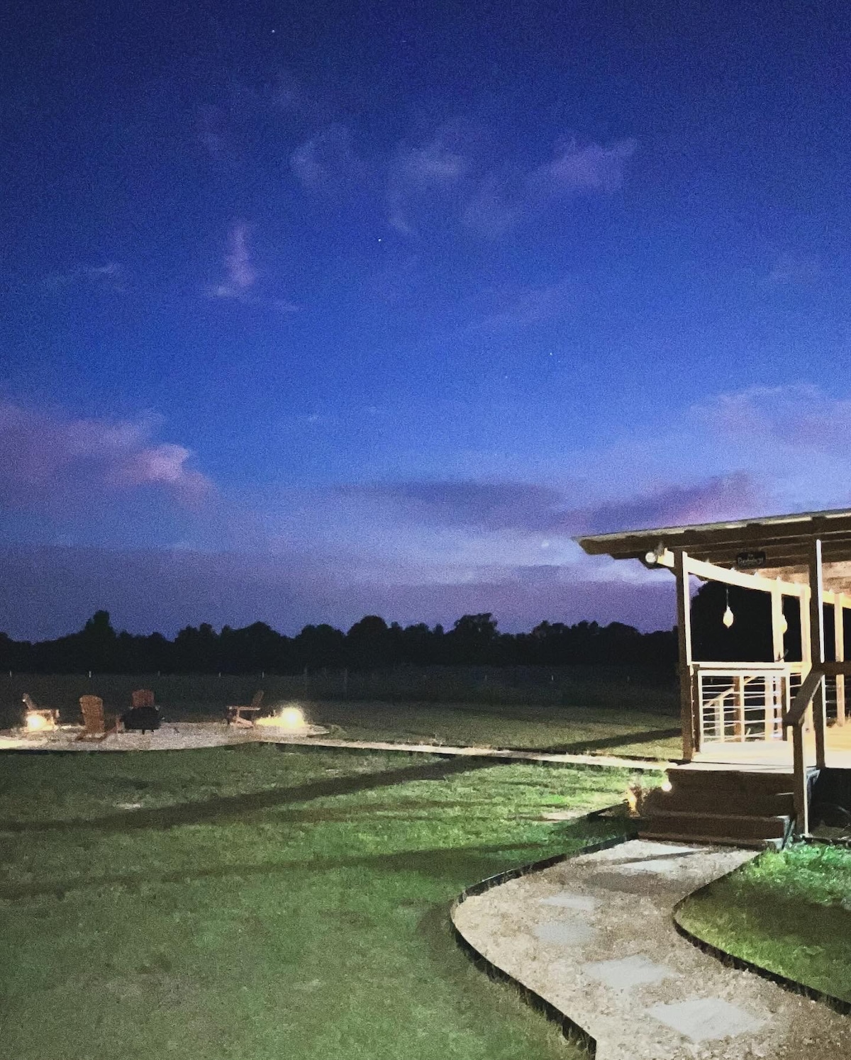 A serene evening scene is captured, showcasing a pathway leading to a cozy porch under a twilight sky. Soft clouds reflect hues of blue and purple, while a fire pit area with seating is visible in the distance, surrounded by expansive green land.