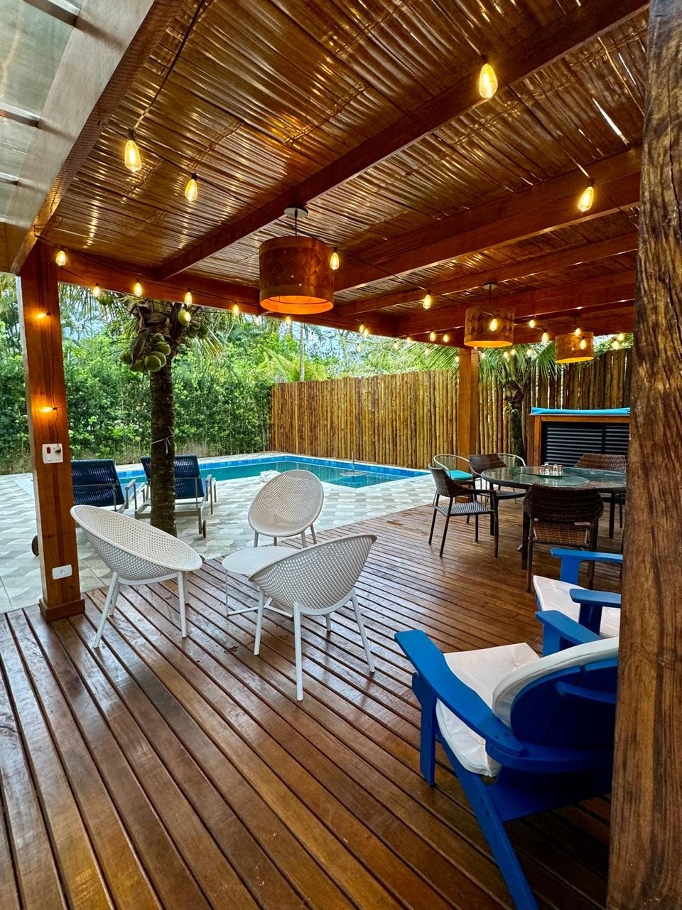A covered outdoor seating area features wooden decking with modern white chairs and a blue lounge chair. A glass-top dining table is surrounded by dark wicker chairs. Soft lighting is provided by hanging lamps, while a private pool is visible in the background, surrounded by lush greenery.