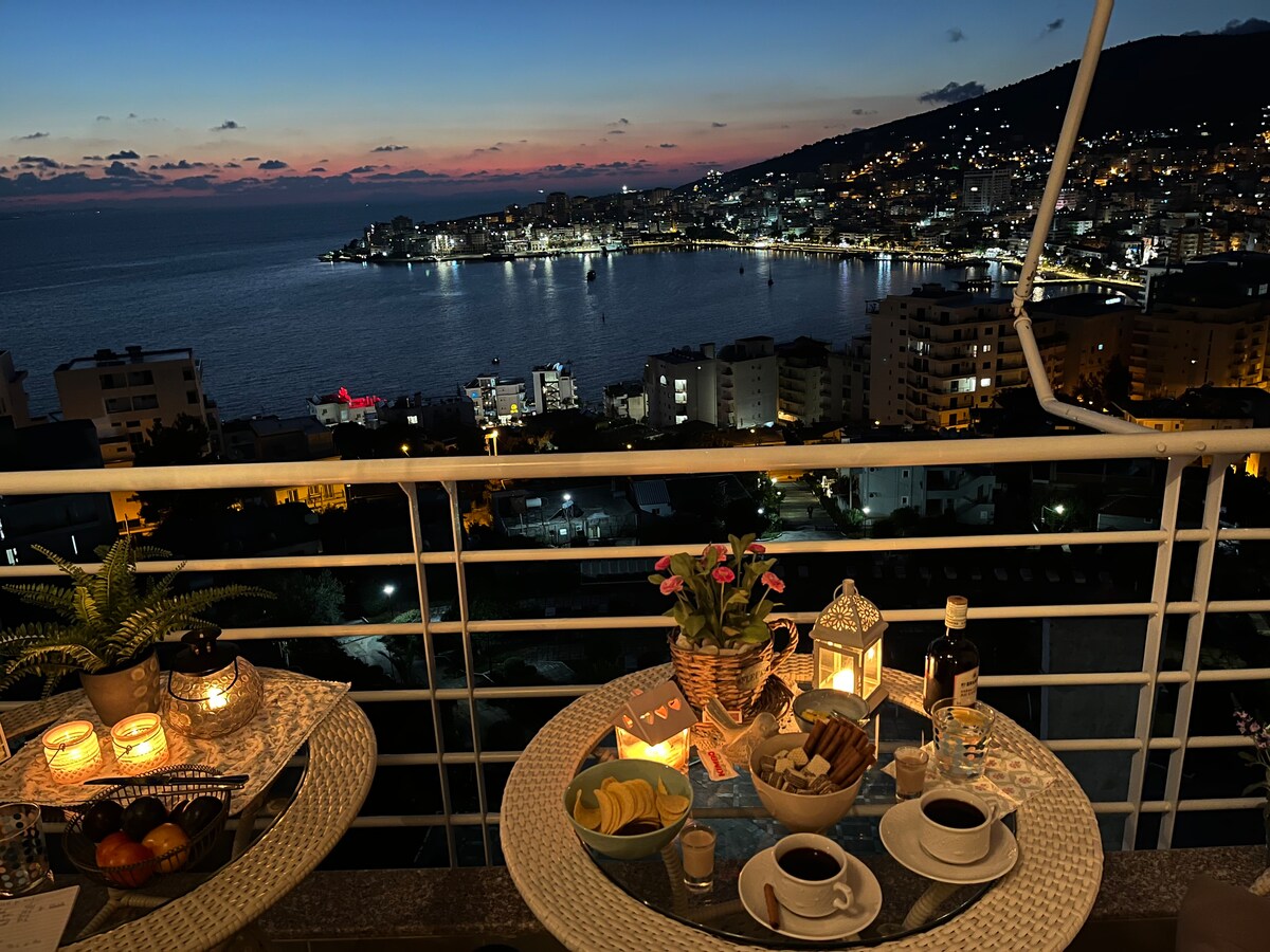 An evening view showcases a beautifully set table with a floral centerpiece and warm candlelight. Scenic sights of Sarandë and the coastline are visible, as the sky transitions to twilight, reflecting soft hues on the water. The balcony offers an inviting space for relaxation.