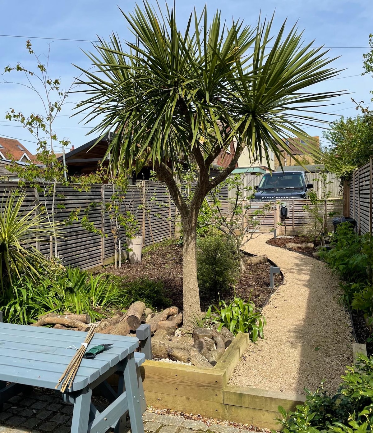 A mature garden path is bordered by landscaping featuring various plants and trees, including a tall palm-like tree. A rustic table sits in the foreground, with a gravel walkway leading through the manicured greenery. A vehicle is visible at the garden's end.