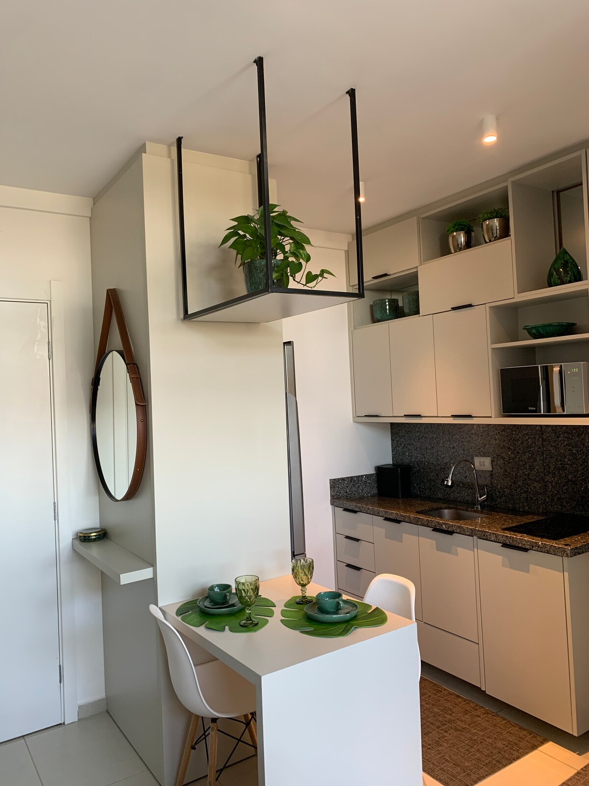 A compact kitchen area features modern cabinetry and a dark granite countertop. A table for two is set with decorative plates and glasses, surrounded by stylish chairs. Above, a hanging plant adds a touch of greenery, complemented by a circular mirror on the wall.