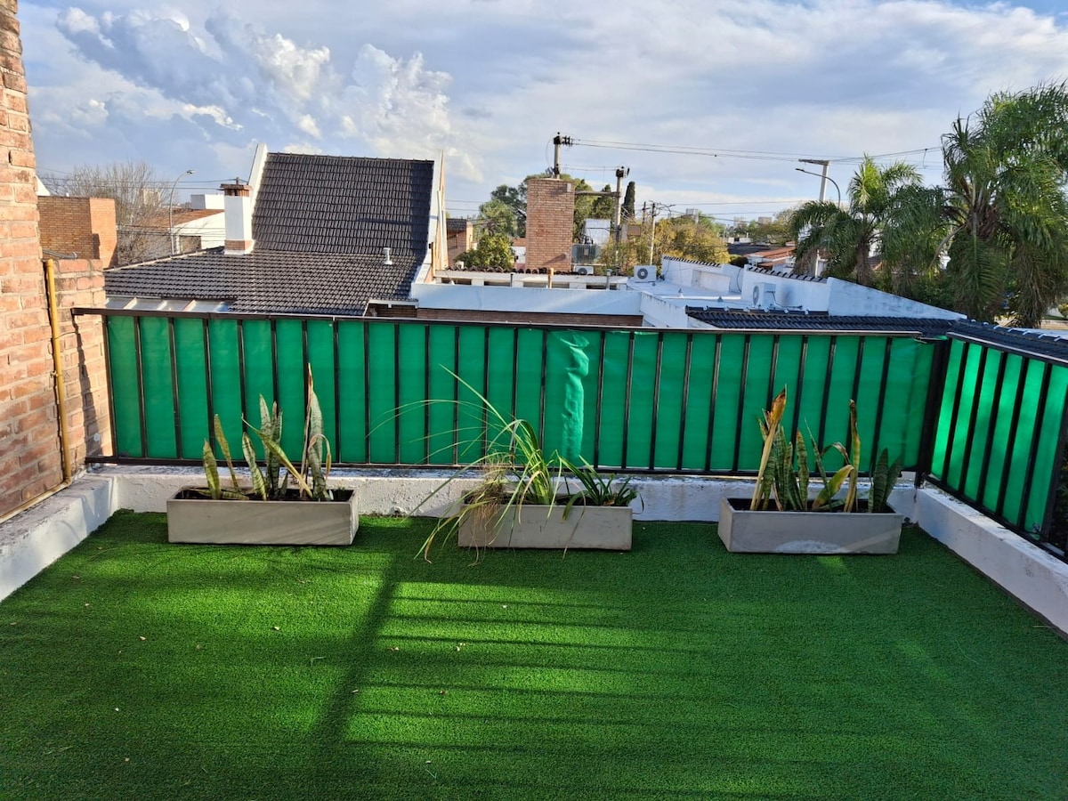 The balcony showcases artificial grass and planter boxes with various plants. Green privacy screens border the space, while the surrounding rooftops and trees are visible in the background. Natural light creates shadows, enhancing the outdoor atmosphere.