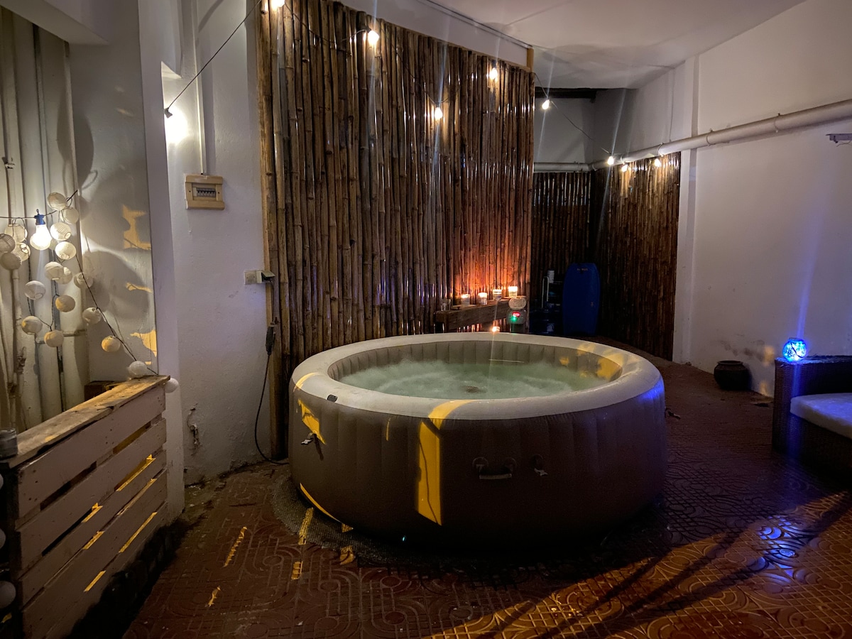 A circular jacuzzi is positioned in a softly lit area, surrounded by a bamboo wall. The water is gently bubbling, creating a tranquil atmosphere. Soft lighting reflects off the surface, enhancing the inviting space for relaxation.