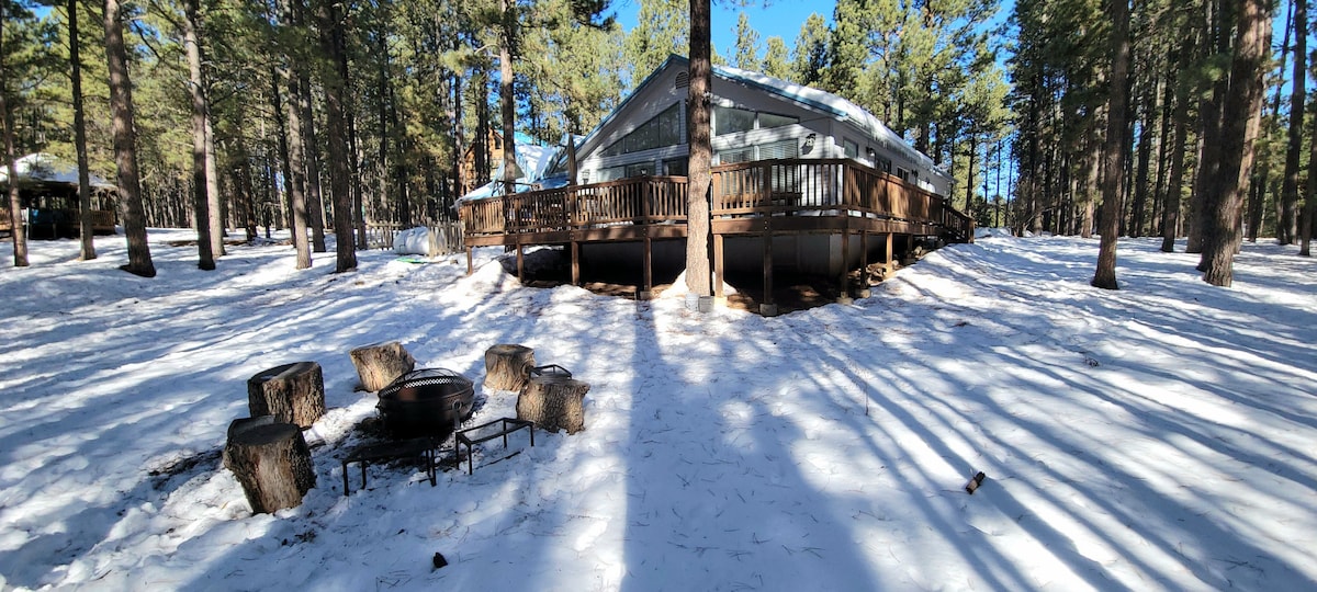 The exterior of Dubbie's Lodge is surrounded by tall pine trees and a snow-covered landscape, creating a serene winter setting. A wooden deck extends around the lodge, with a designated fire pit area featuring several seating logs.