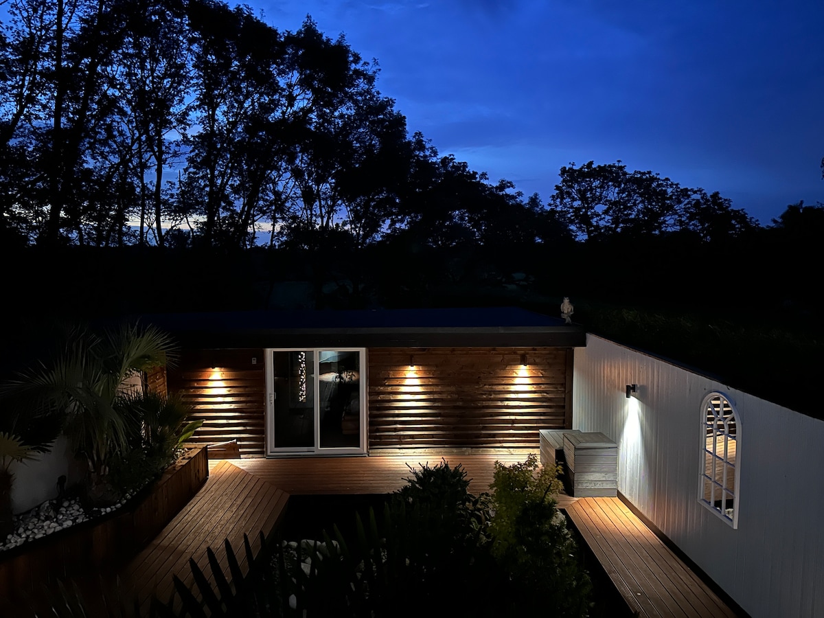The cabin is illuminated under a twilight sky, showcasing a welcoming outdoor area with timber decking. Lush greenery surrounds the space, while soft lighting highlights the entrance. A seating area is positioned on the deck, creating a serene environment for relaxation.