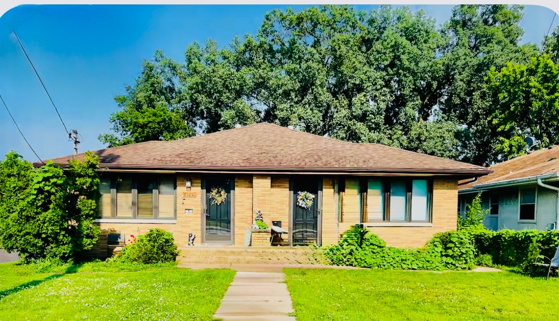 A single-story brick duplex is captured from the front, framed by lush greenery and trees. The entrance features a pathway leading to a porch with decor that includes flower wreaths on the windows, enhancing the inviting exterior.