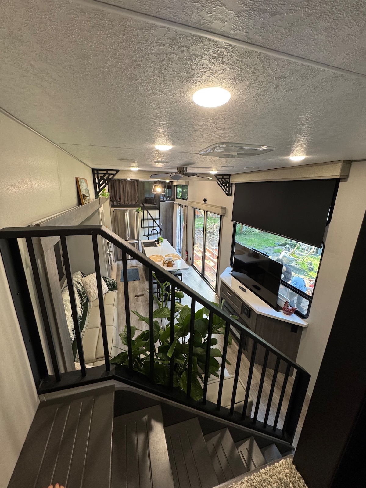 A view from the staircase captures the open living area below, featuring large windows that invite natural light. An indoor plant adds a touch of greenery beside a television. A dining table with seating is visible in the background, blending comfort with a modern aesthetic.