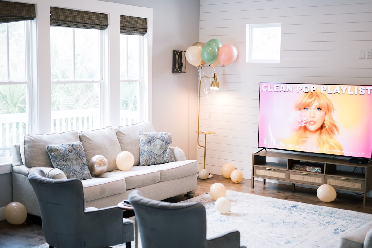 A cozy living area features a light-colored sofa adorned with blue and white patterned cushions. Two dark chairs provide additional seating. A large TV displays a colorful image, while balloons in soft tones are scattered around the room, adding a festive touch. Natural light floods through multiple windows.