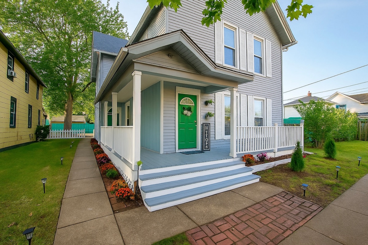 The exterior of the duplex is captured, showcasing a welcoming porch with a bright green door framed by flower decorations. A well-maintained lawn features colorful flower beds and concrete pathways leading up to the entrance.