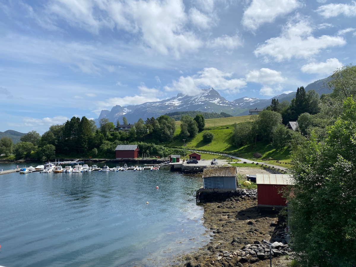 A serene coastal view captures the still water reflecting the sky. Small boats are anchored along the shore, with red sheds dotting the landscape. Lush greenery rises in the background, leading to distant mountains capped with snow under a partly cloudy sky.