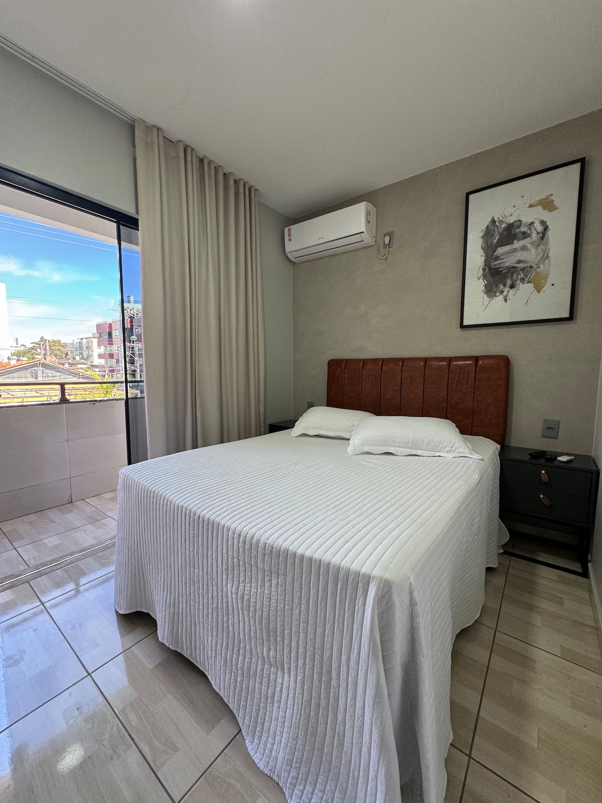 A cozy sleeping area features a spacious bed with a white bedspread and soft pillows. Natural light enters through large glass doors that open to a balcony. Air conditioning is mounted on the wall, and a piece of artwork is displayed above a nightstand.