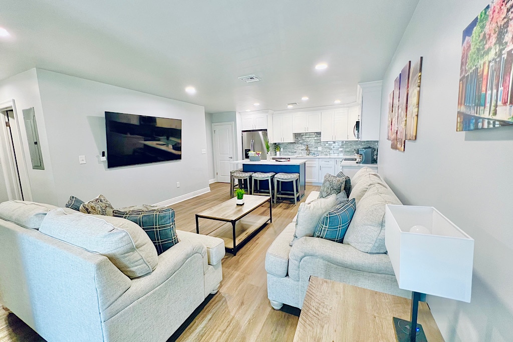 The open living area features a light-colored sofa set with plaid cushions, facing a large flat-screen TV mounted on the wall. A wooden coffee table sits in front of the sofas, and a modern kitchen with bar seating is visible in the background.
