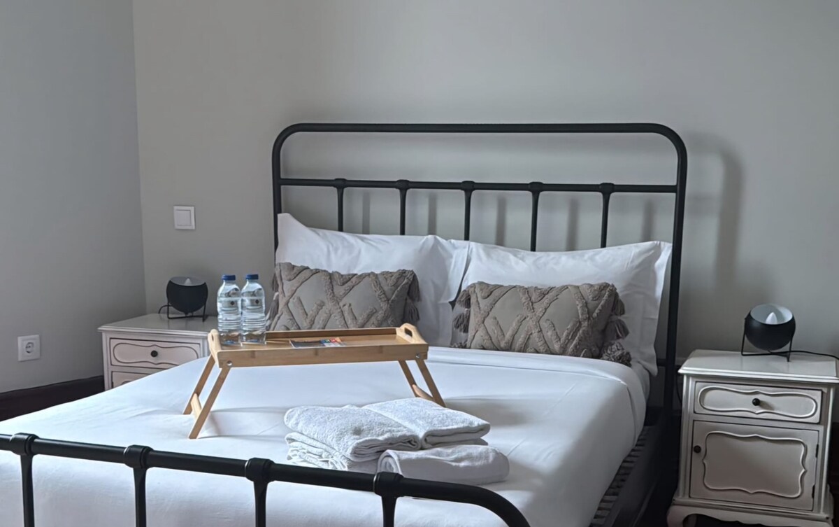 A comfortable bedroom features a metal bed frame with a spacious bed topped with white linens and textured pillows. A small wooden tray holds two water bottles and stationery, while white towels are neatly arranged at the foot of the bed. Nightstands with modern lamps flank the bed.