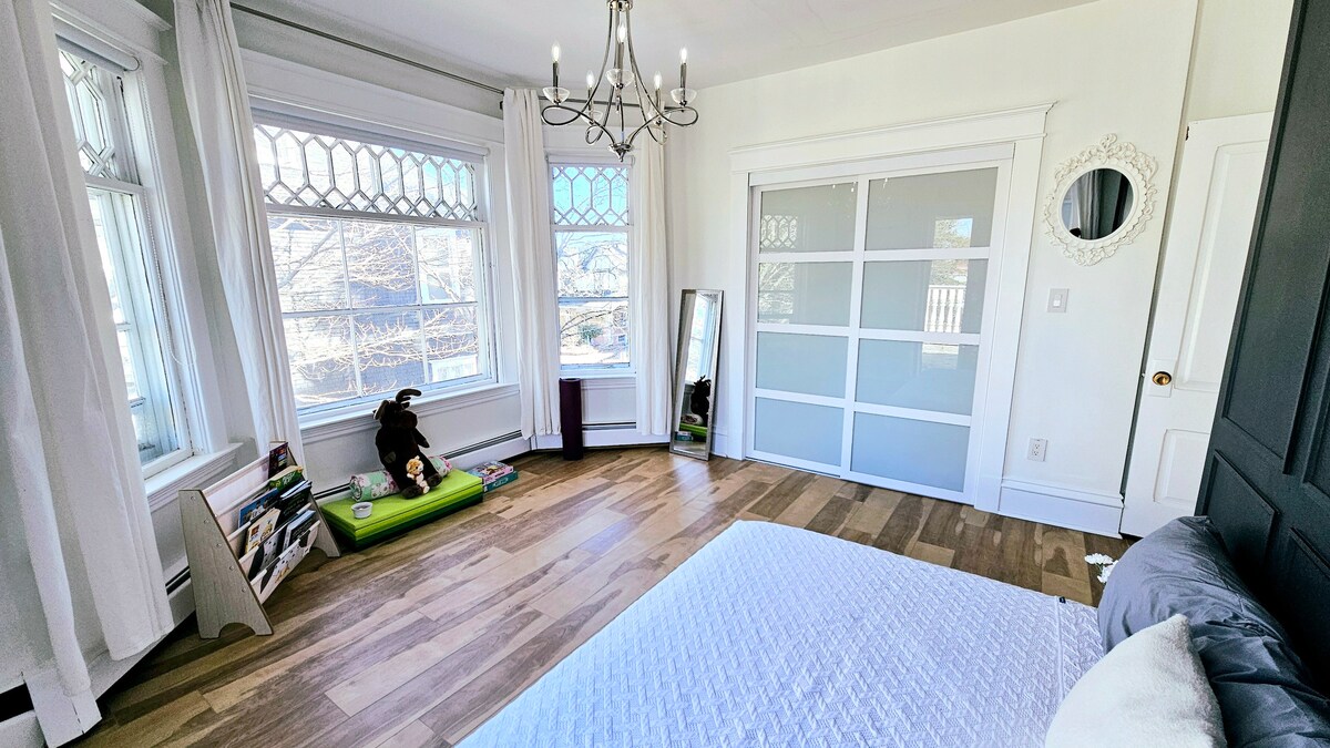 A spacious bedroom features large windows with decorative stained glass, allowing natural light to fill the room. A cozy bed is present, along with a full-length mirror and a sleek sliding door wardrobe. A green seating area with books adds a touch of comfort.