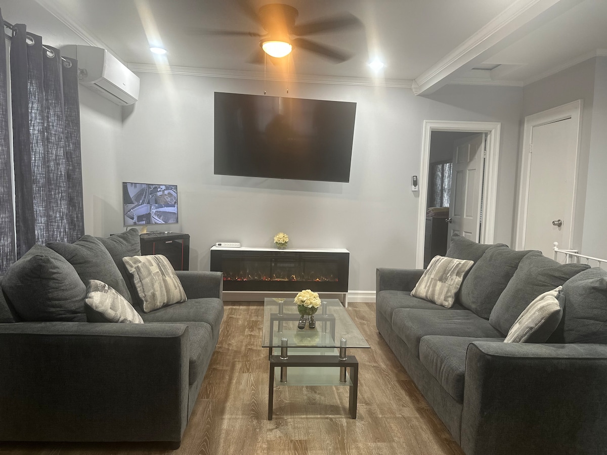 The living room features two dark grey couches, each adorned with decorative pillows. A glass coffee table sits in the center, complemented by a small vase with flowers. A wall-mounted television is positioned above an electric fireplace, and natural light filters through sheer curtains.