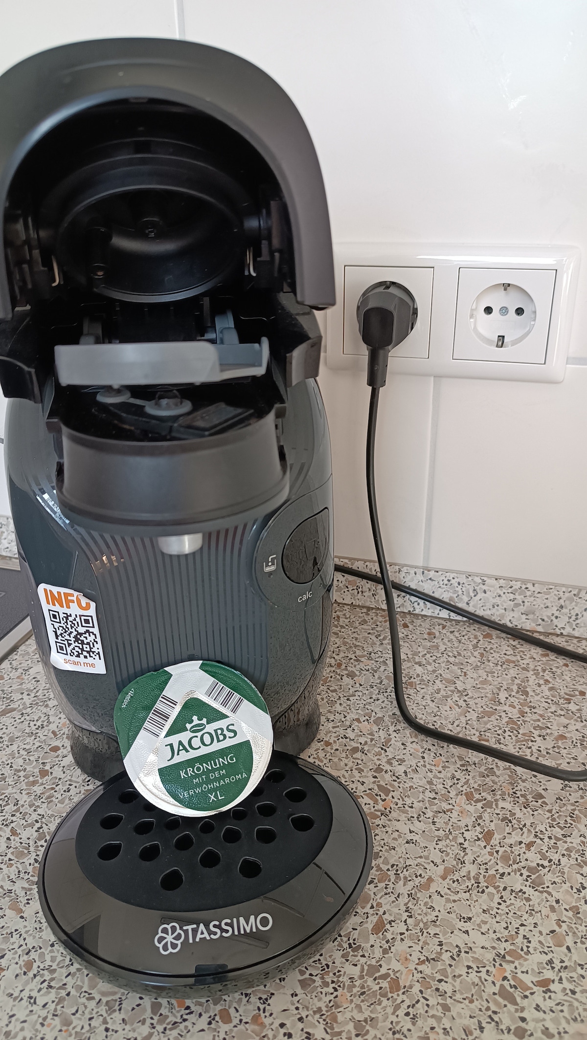 A coffee machine is positioned on a textured countertop, featuring a pod holder with a Jacobs coffee pod. The machine has a sleek design and is connected to a power outlet, indicating accessibility for brewing. The surrounding area includes a light-colored backsplash.