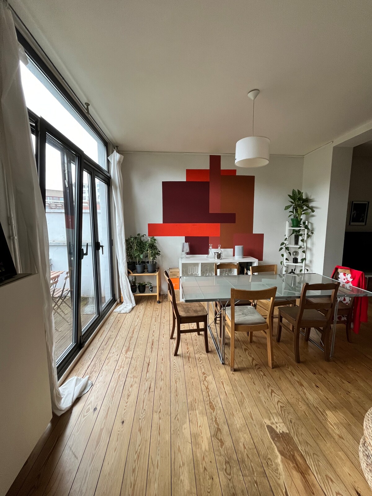 A dining area is present, featuring a rectangular glass table surrounded by wooden chairs. A colorful abstract mural adorns the wall, complemented by potted plants. Large windows allow natural light to fill the space, while light-colored curtains frame the view.