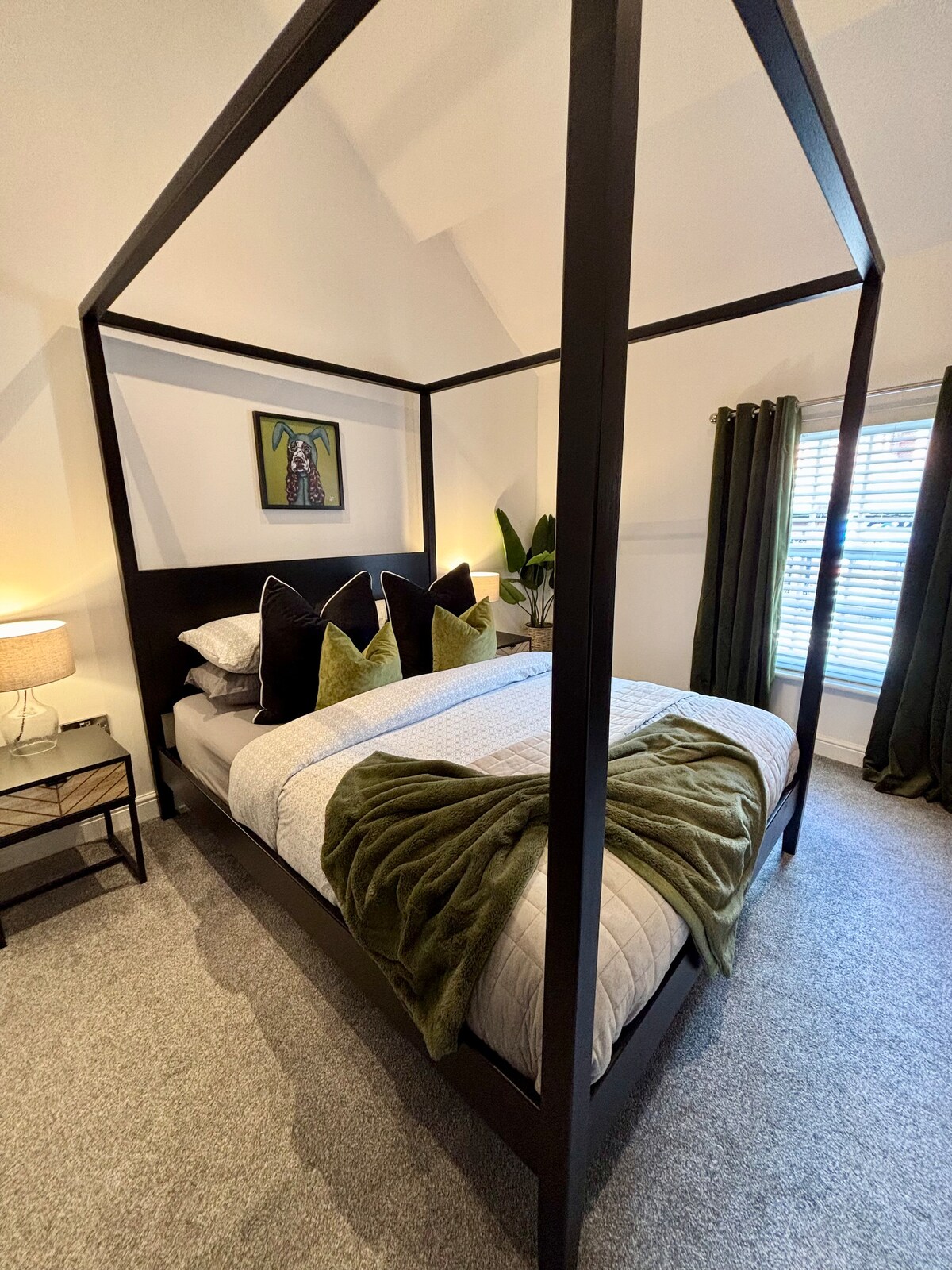 A stylish bedroom is highlighted by a four-poster bed with plush green accent pillows and a textured throw draped at the foot. Soft lighting from a bedside lamp complements the subtle color palette, while curtains frame the window, allowing natural light to filter in.