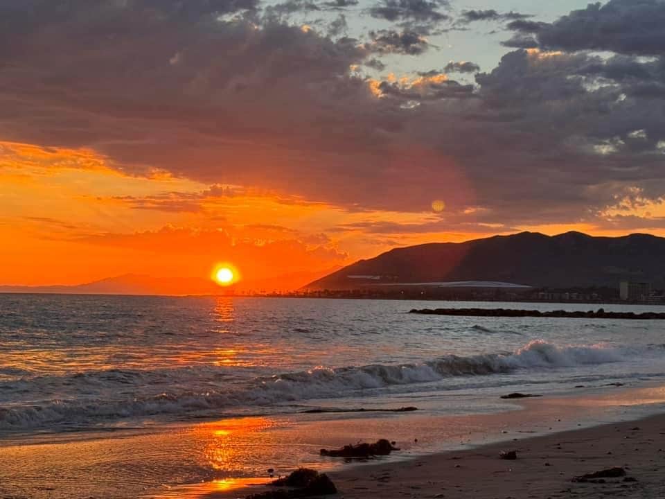 A vibrant sunset casts warm orange and yellow hues over the ocean, reflecting on the gentle waves. The silhouette of distant mountains and a coastal structure is visible against the colorful sky, creating a serene evening scene by the beach.