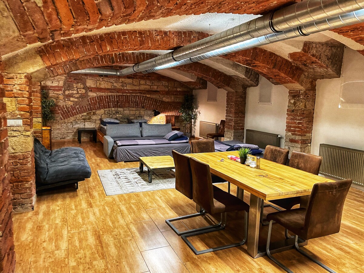 The spacious room features exposed brick walls and vaulted ceilings, enhancing its unique charm. A dining table accommodates several chairs, while multiple beds provide ample sleeping arrangements. A soft couch invites relaxation, and natural light filters through the windows, creating a warm ambiance.