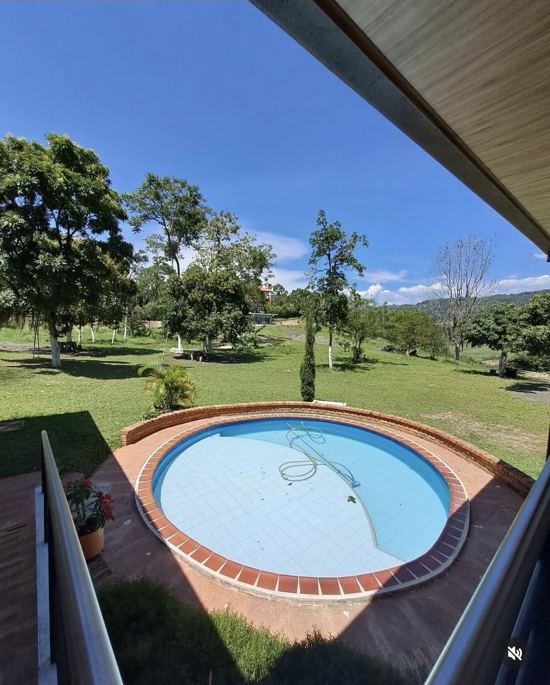A small circular pool is situated amidst a green lawn, surrounded by trees and open space. The clear blue water reflects the sky, while the beige tiled edge complements the natural surroundings. The scene conveys a serene outdoor atmosphere.
